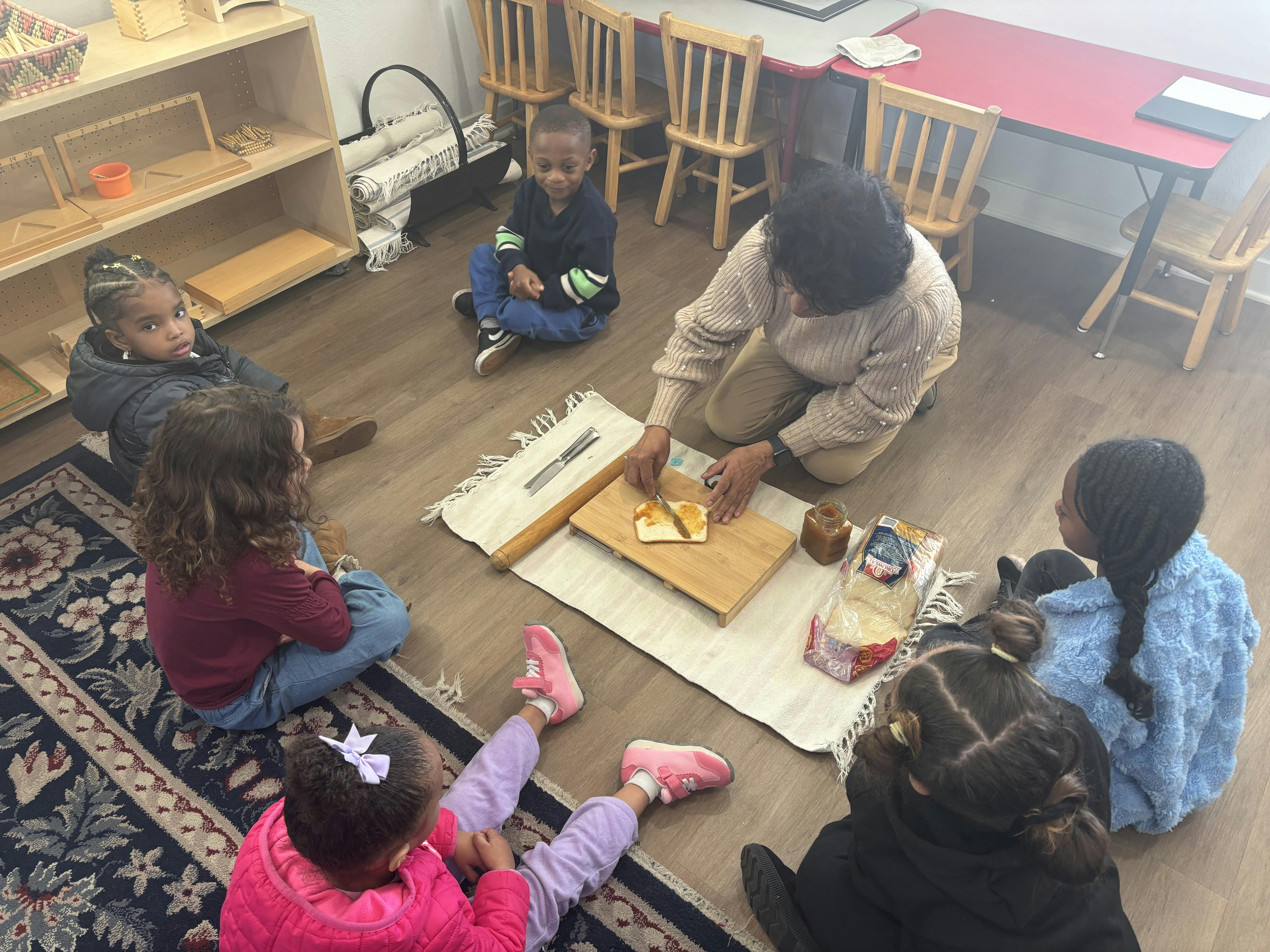 Adult person spreading jam on toast with children gathered around in a classroom