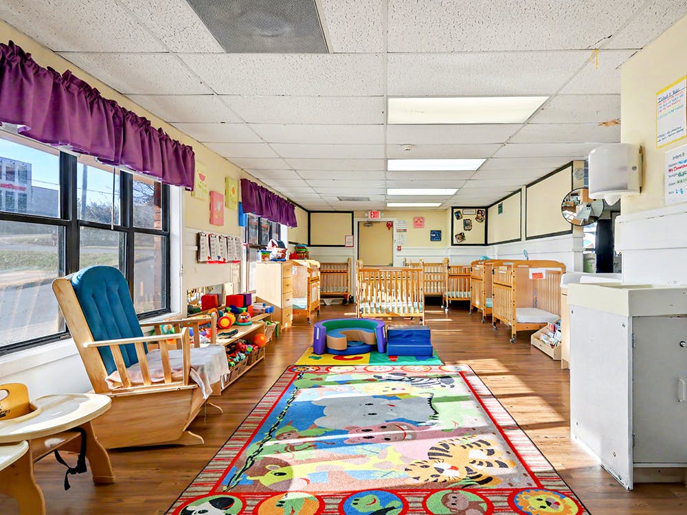 A childcare room with a colorful play rug, wooden cribs, and a rocking chair