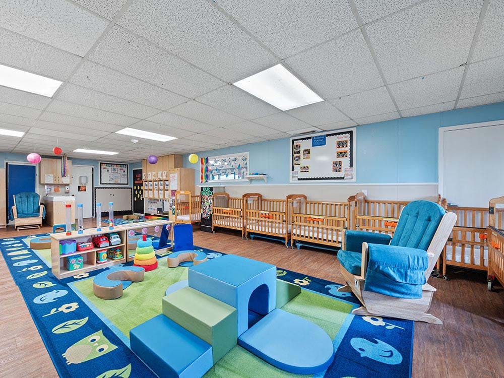 A colorful childcare classroom with cribs, play mats, and a rocking chair