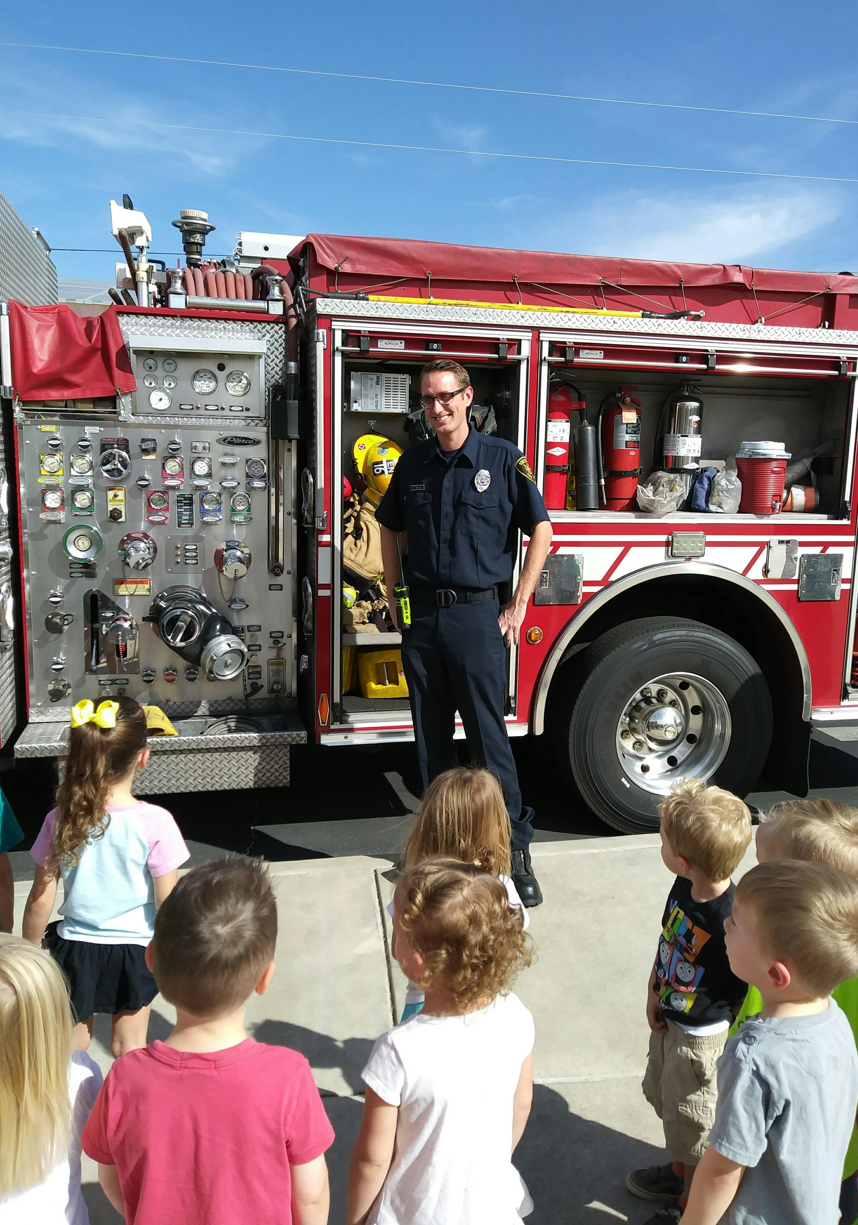 Firefighter standing in front of a fire truck, speaking to a group of children