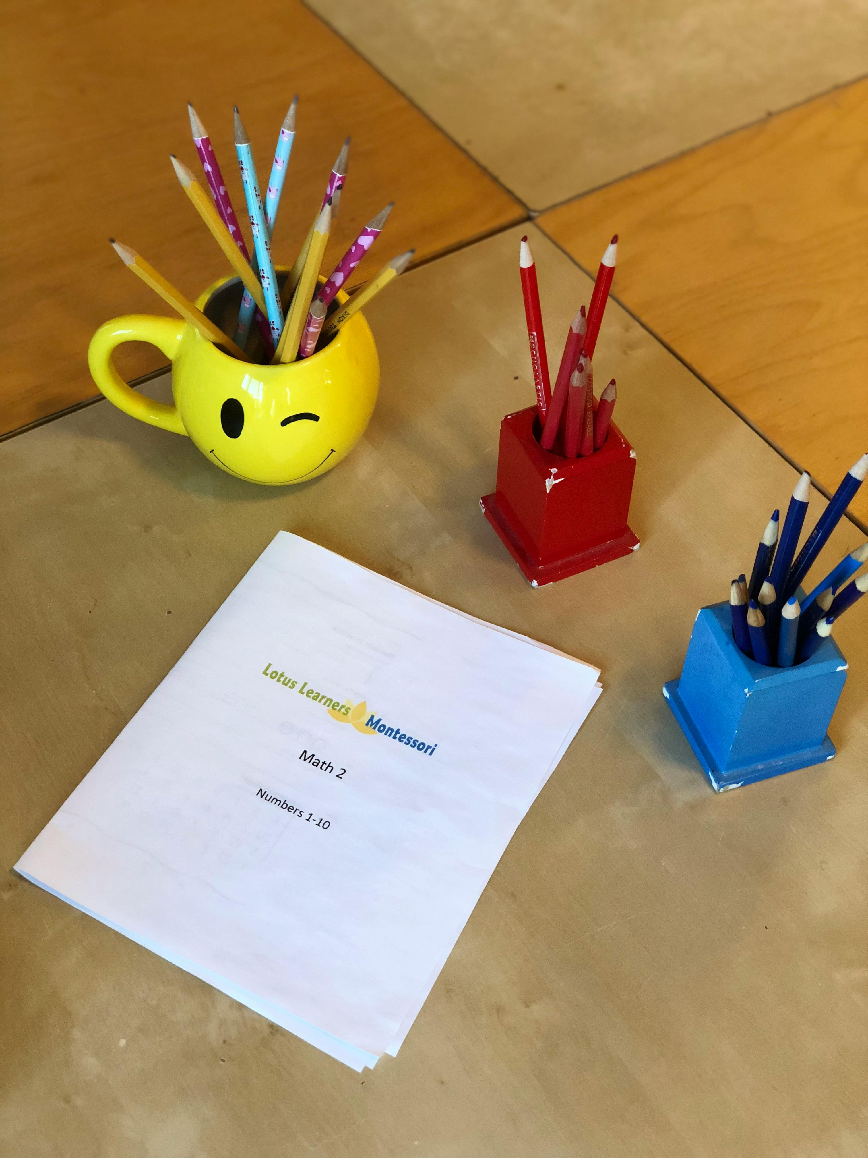 Pencils in a yellow winking face mug, red holder, and blue holder next to a document