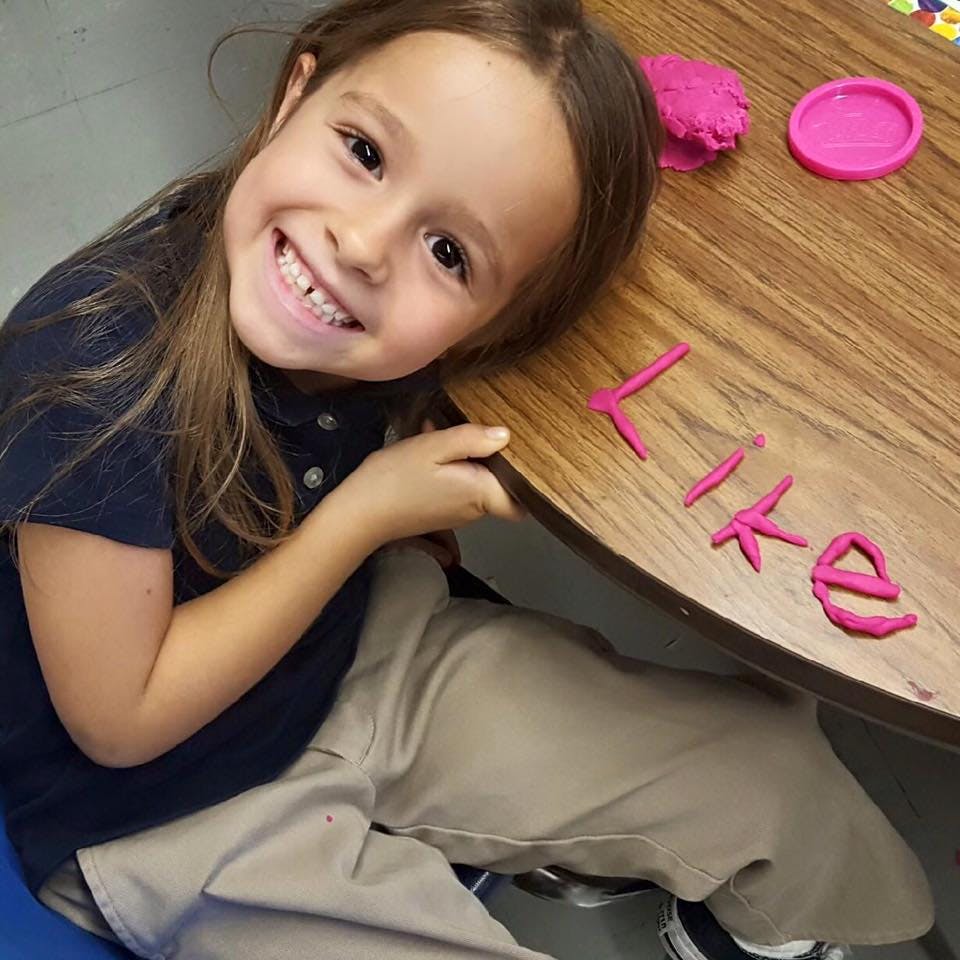 Child smiling at a desk with the word "like" spelled out in pink play-doh