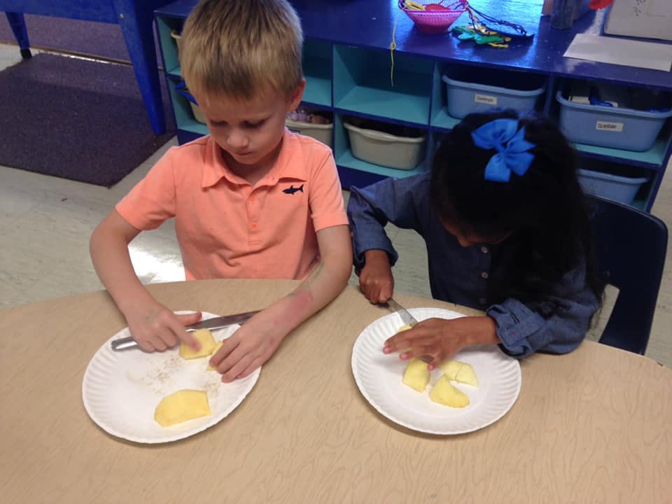 Two children cutting fruit on plates at a table