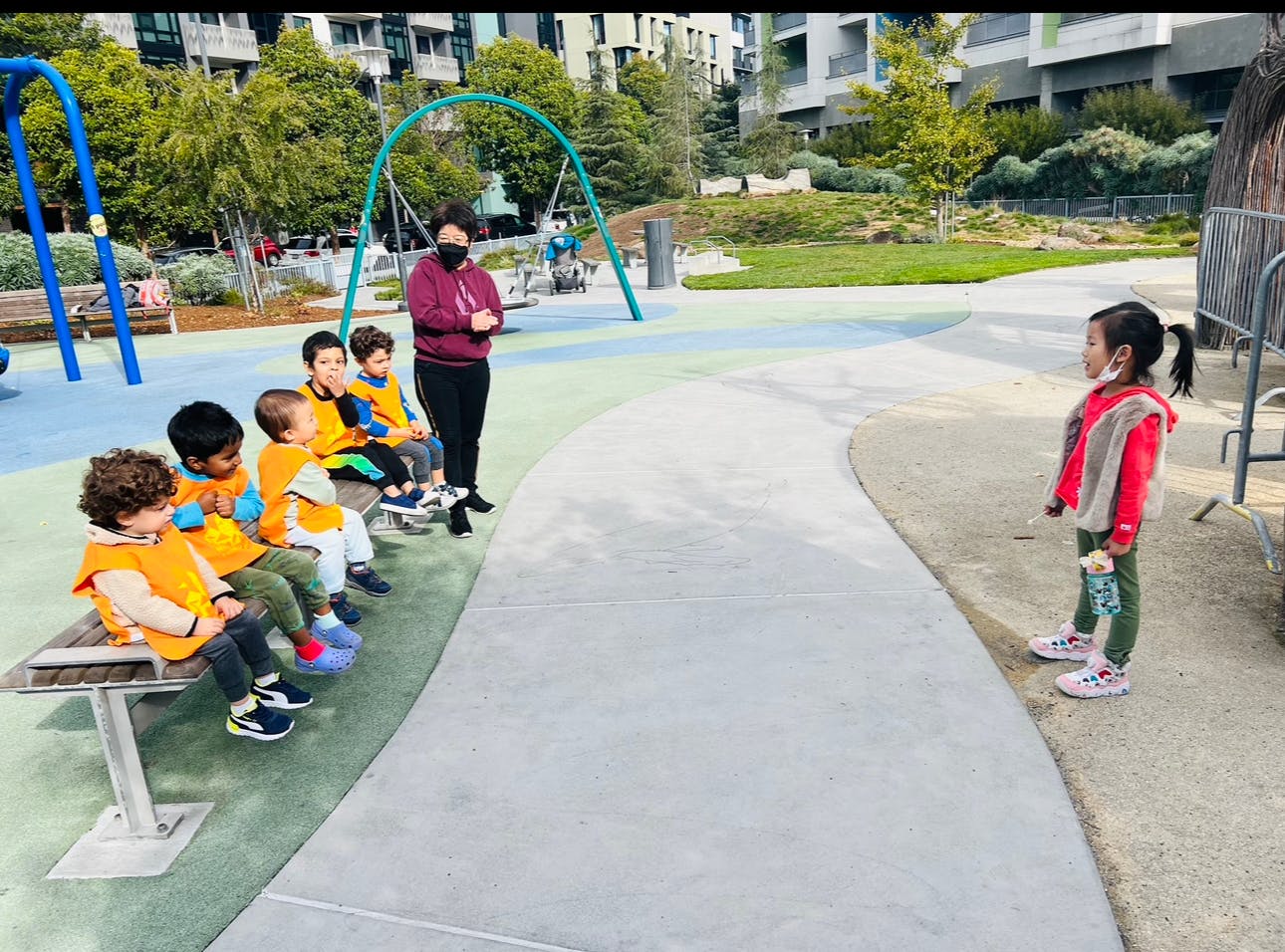 Person in mask with children on a bench and one child standing on a playground