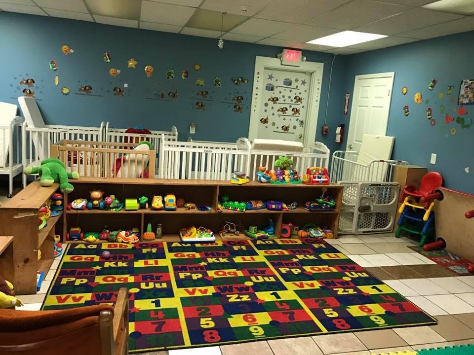 Daycare room with cribs, shelves of toys, and an alphabet rug