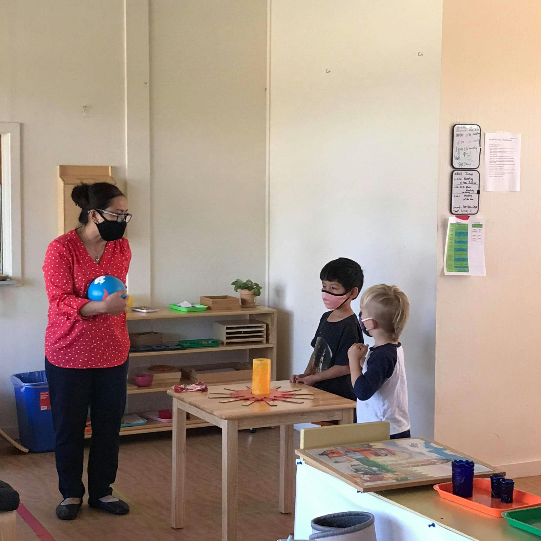Person in mask holding a globe, two children in masks standing by a table