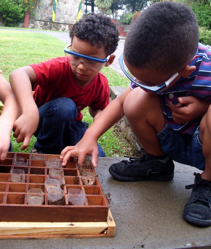 Two children wearing safety glasses examining items in a wooden tray