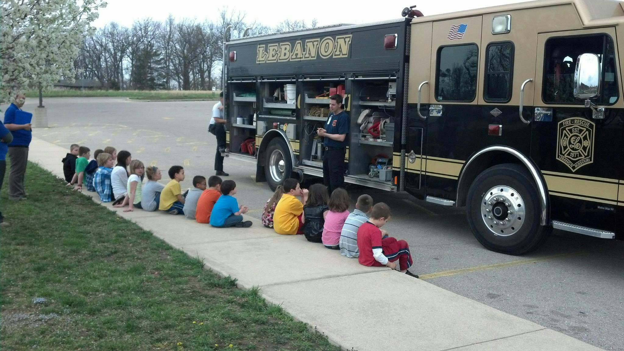 Children sitting on a sidewalk next to a fire truck with a person standing by the truck