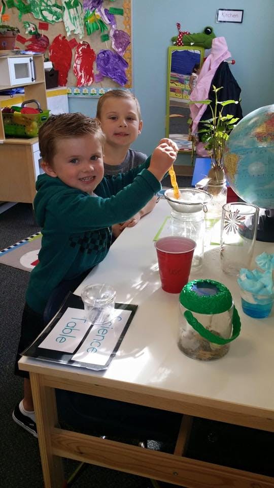 Two children at a science table, one pouring from a spoon into a funnel