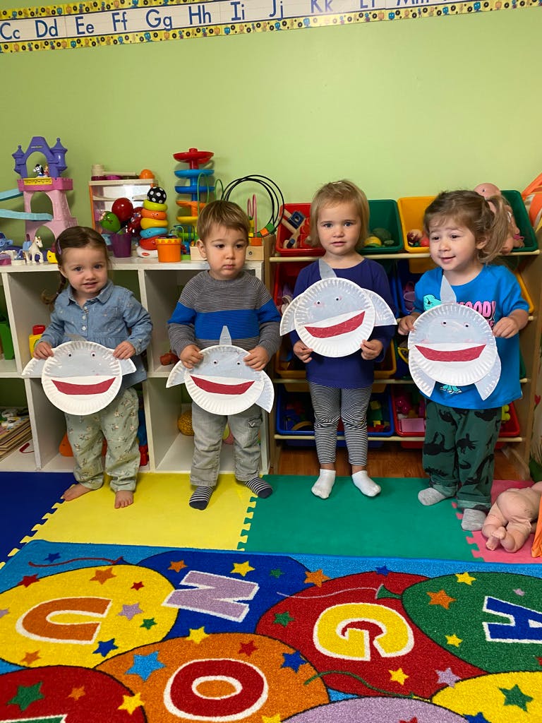 Four children holding paper plate shark crafts in a classroom