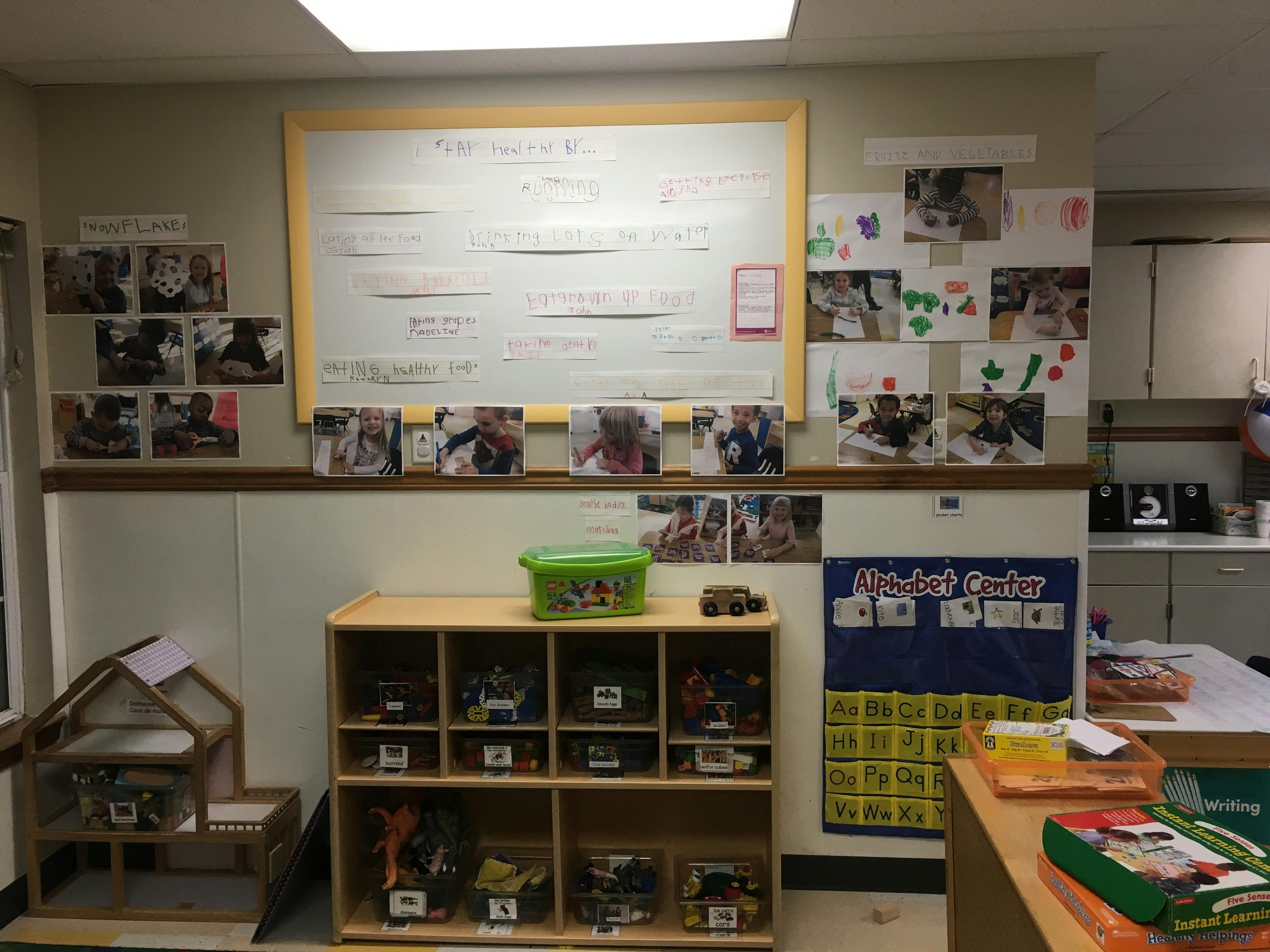 Classroom wall with children's artwork and photos, storage shelves with toys, and an alphabet chart