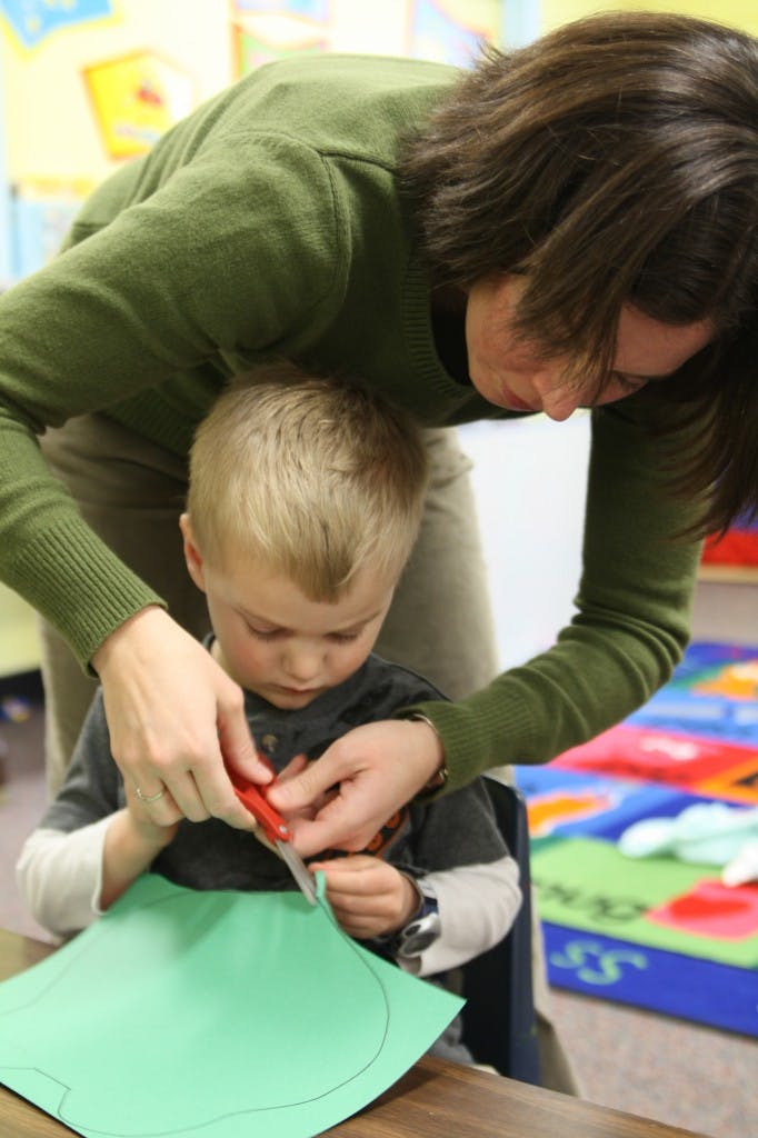 Person helping a child cut green paper with scissors