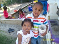 Two children standing, one holding a cup, with other children in a sandbox