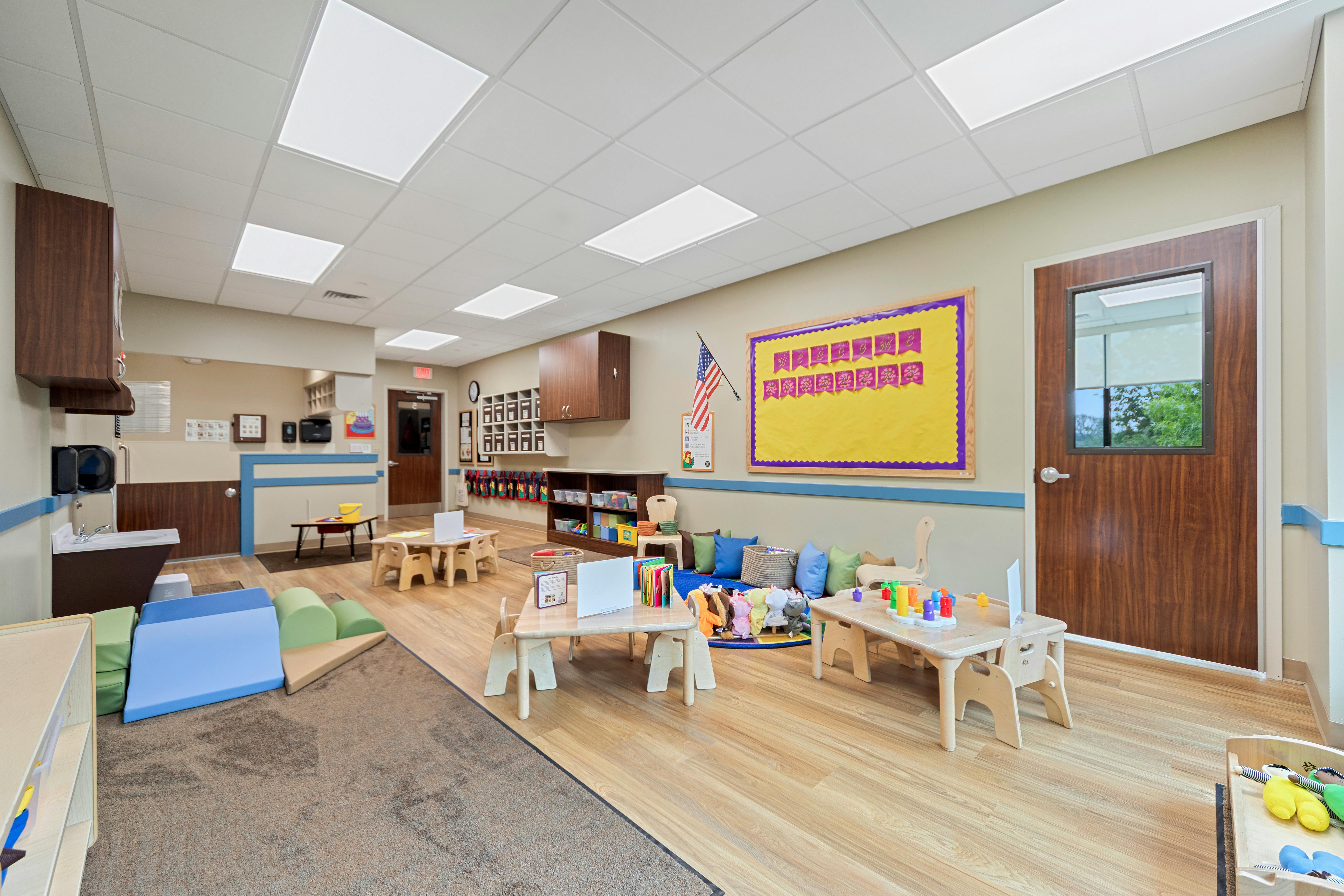A brightly lit classroom with small tables, chairs, play mats, and a bulletin board