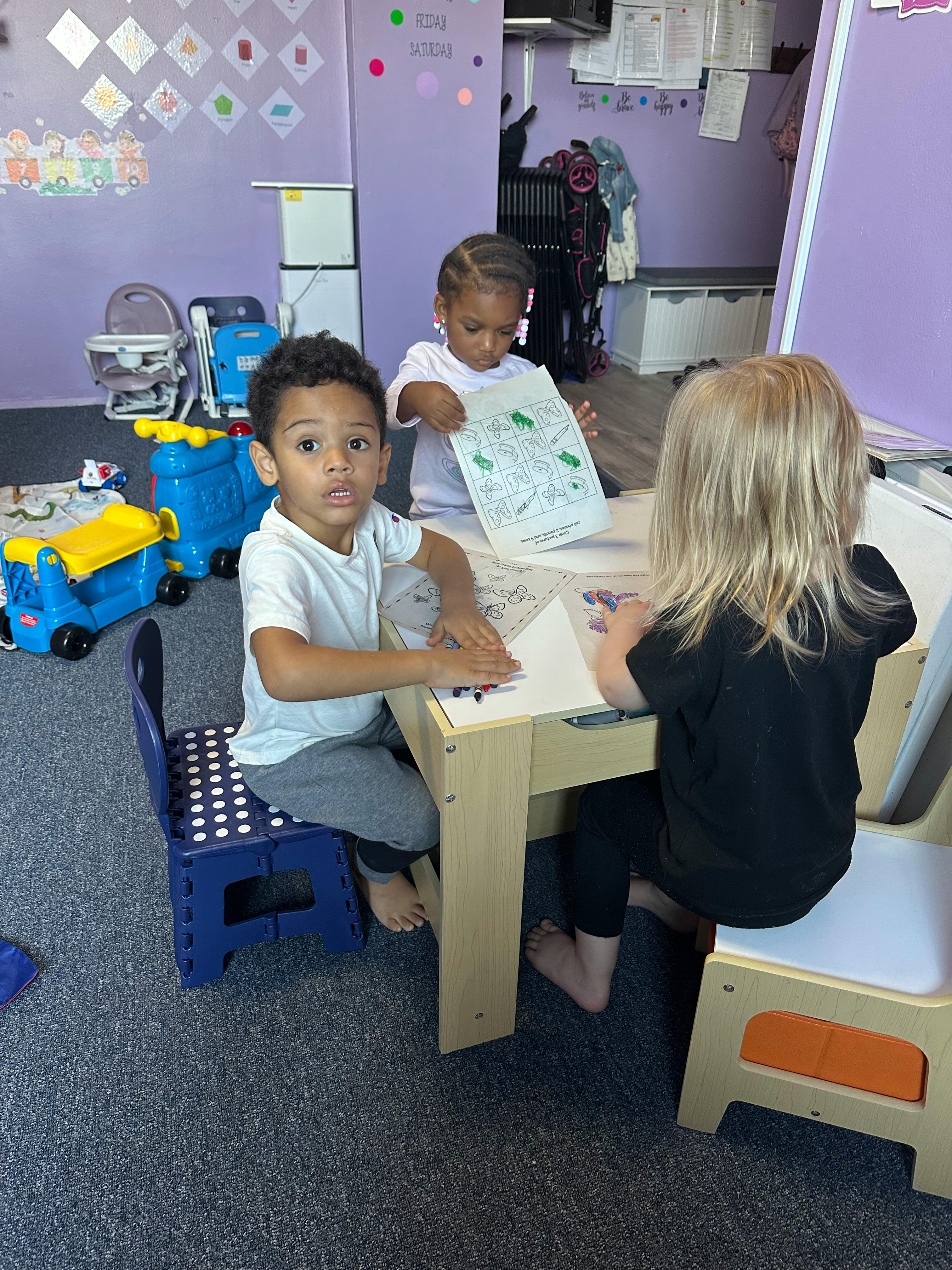 Three children sitting at a small table, two drawing and one holding a paper