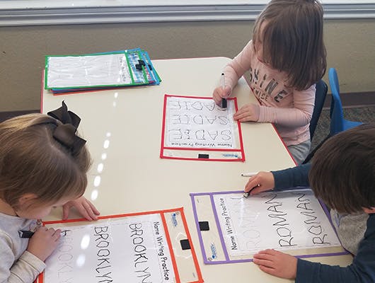 Three children practicing writing names at a table