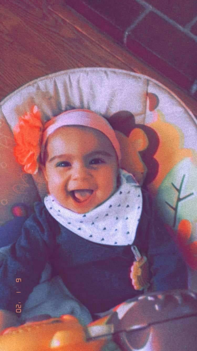 Child in a baby seat with a headband and bib, sticking out tongue