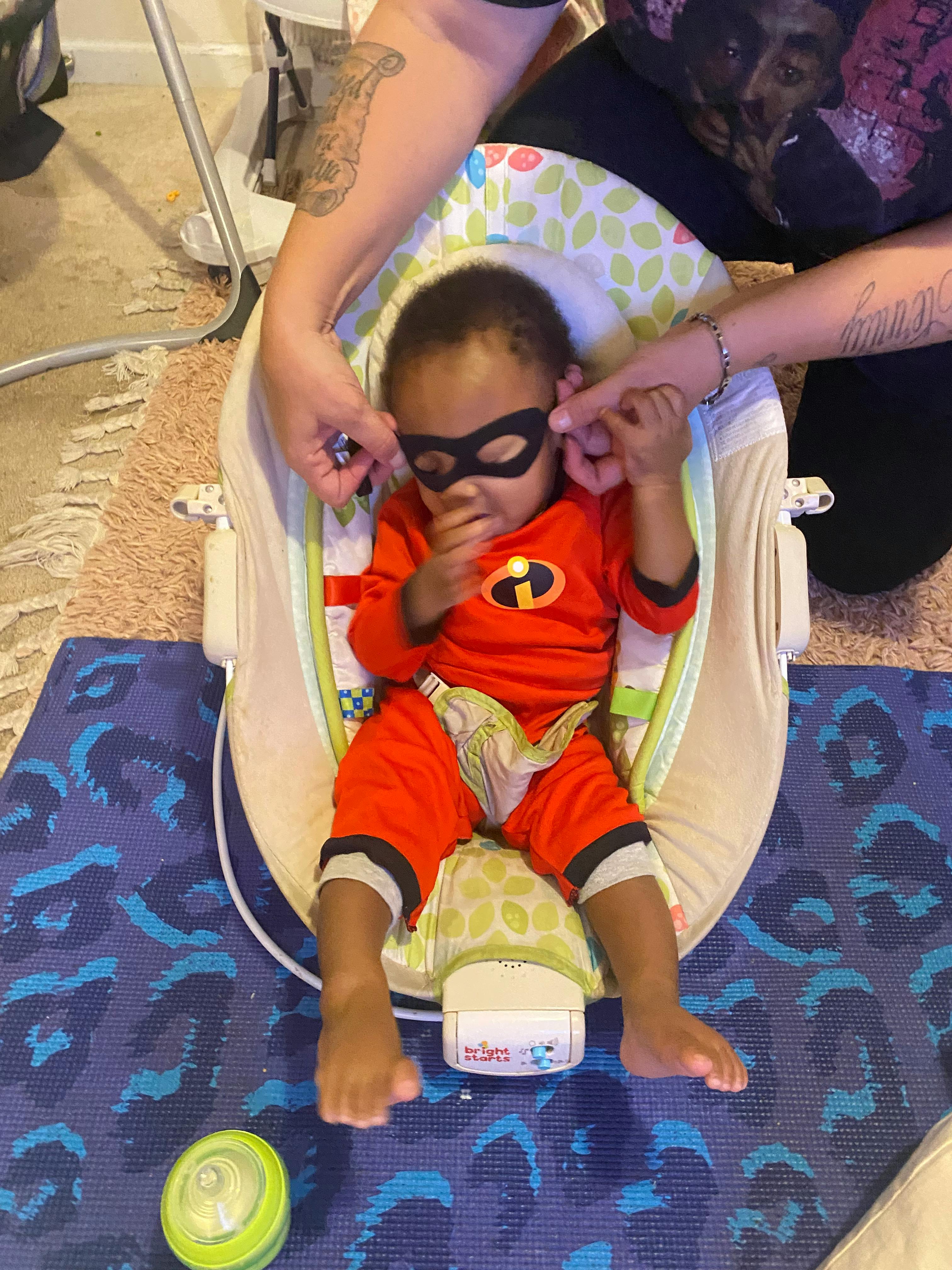 Child in a red superhero costume and black mask sitting in a baby bouncer