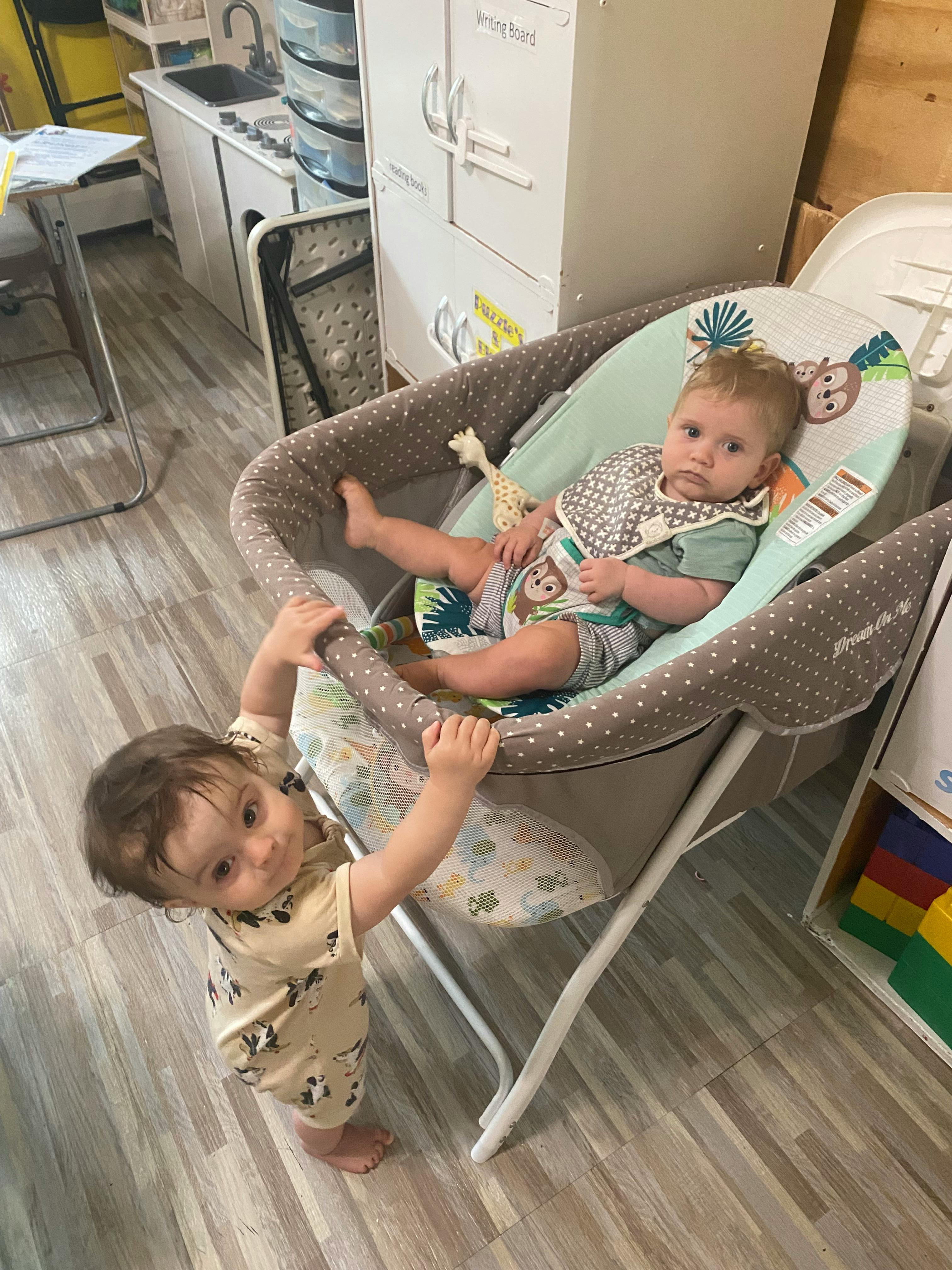 Child sitting in a baby bouncer next to a standing child