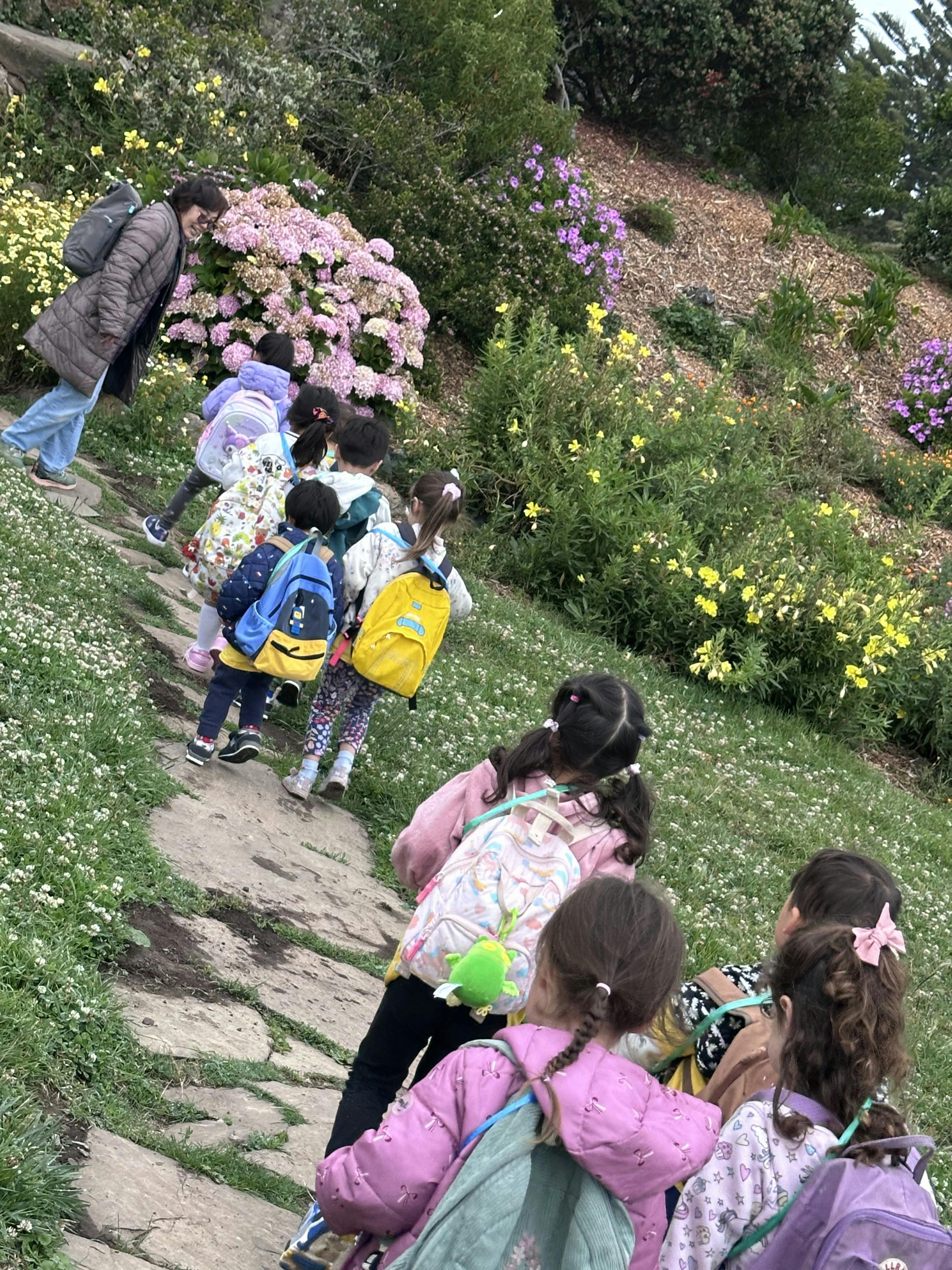 A line of children wearing backpacks walking along a path in a flower garden