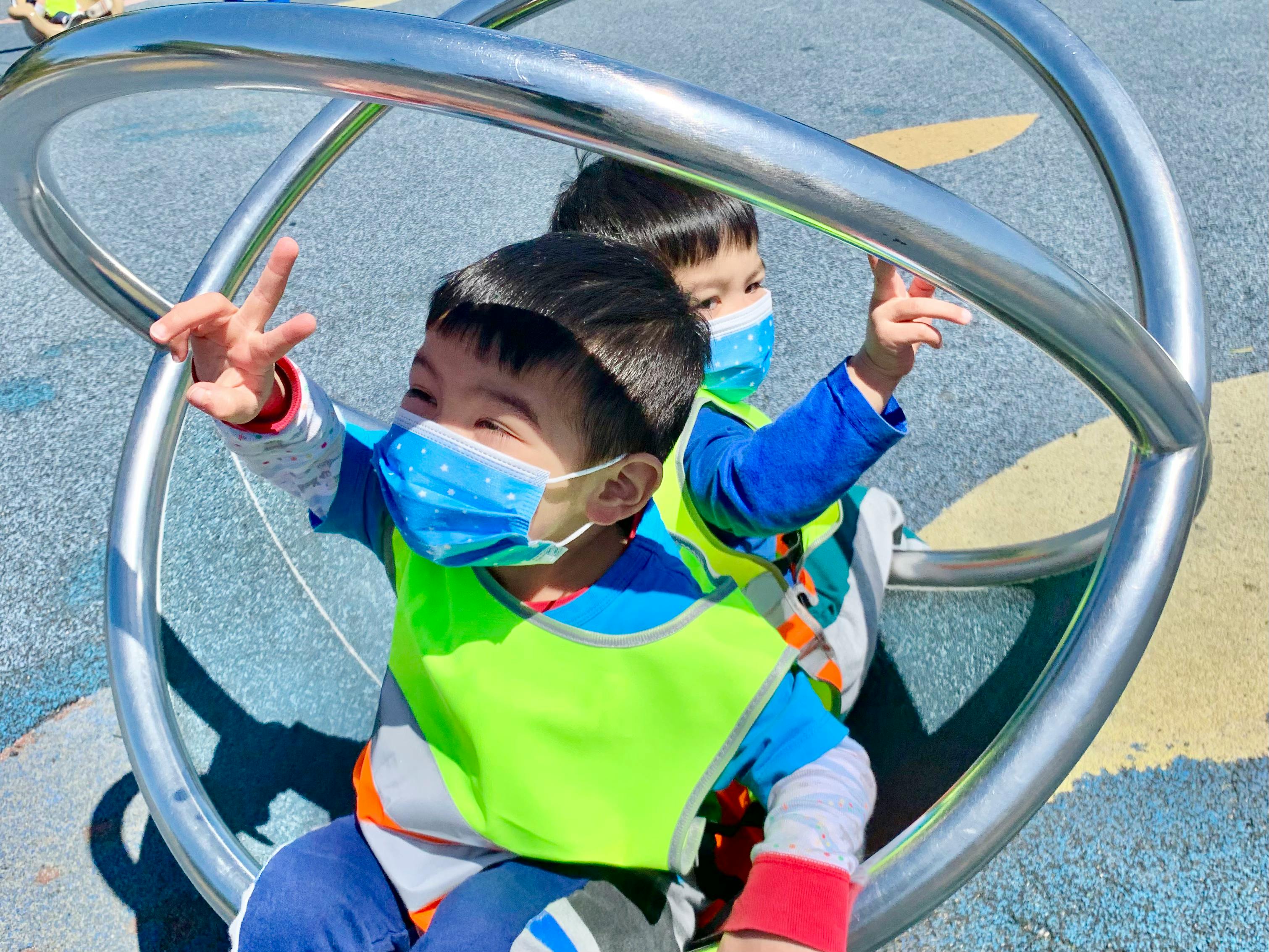Two children wearing masks on a playground spinner