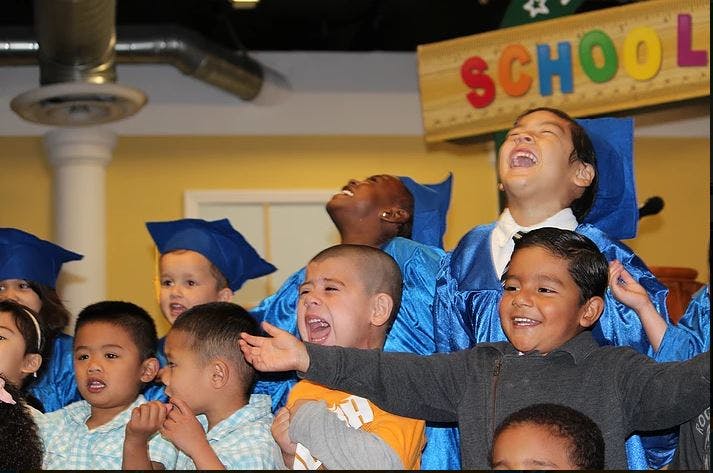 Children in graduation caps and gowns, some laughing, with a "SCHOOL" sign