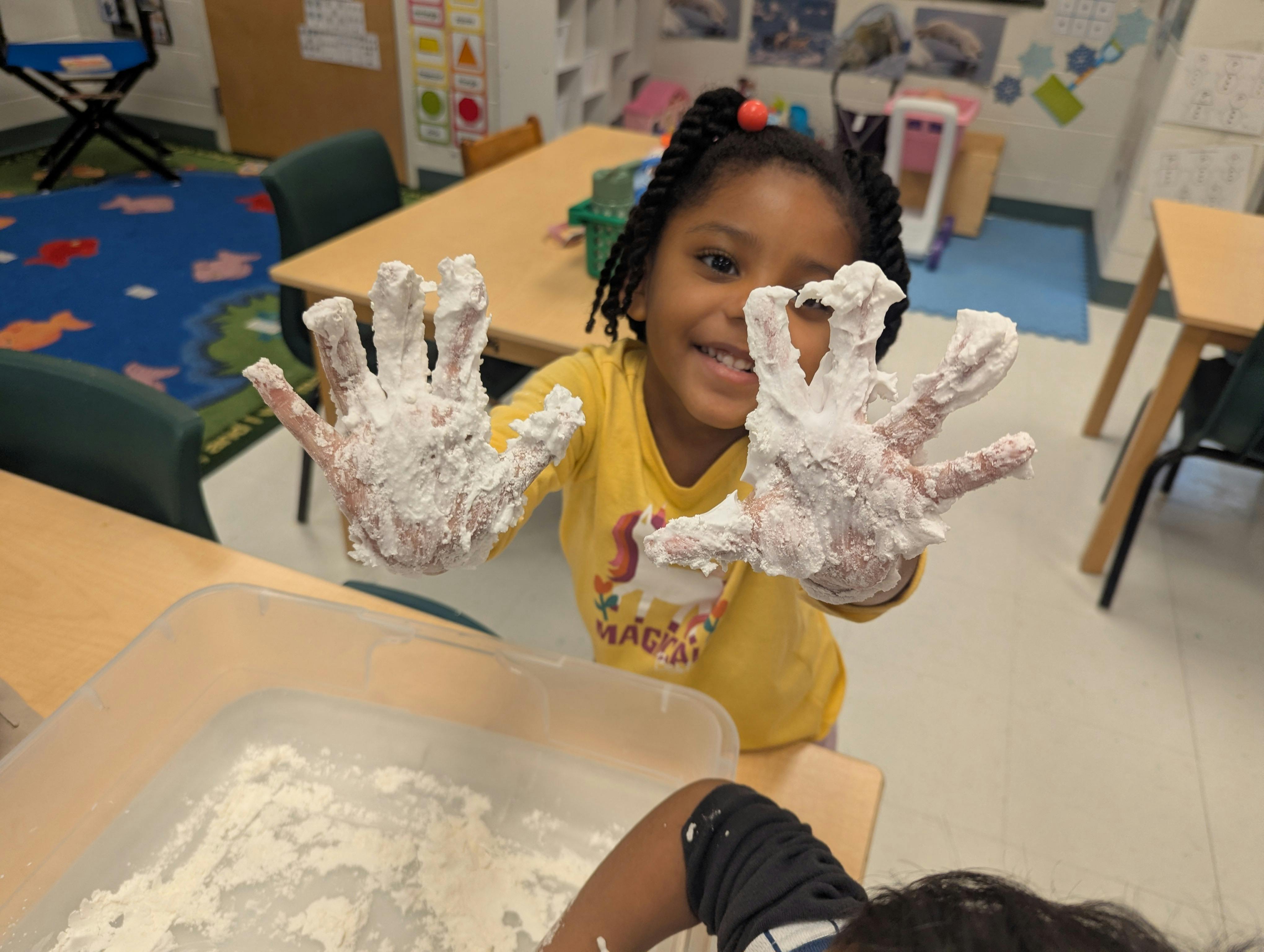 Child in a classroom holding up hands covered in white powder