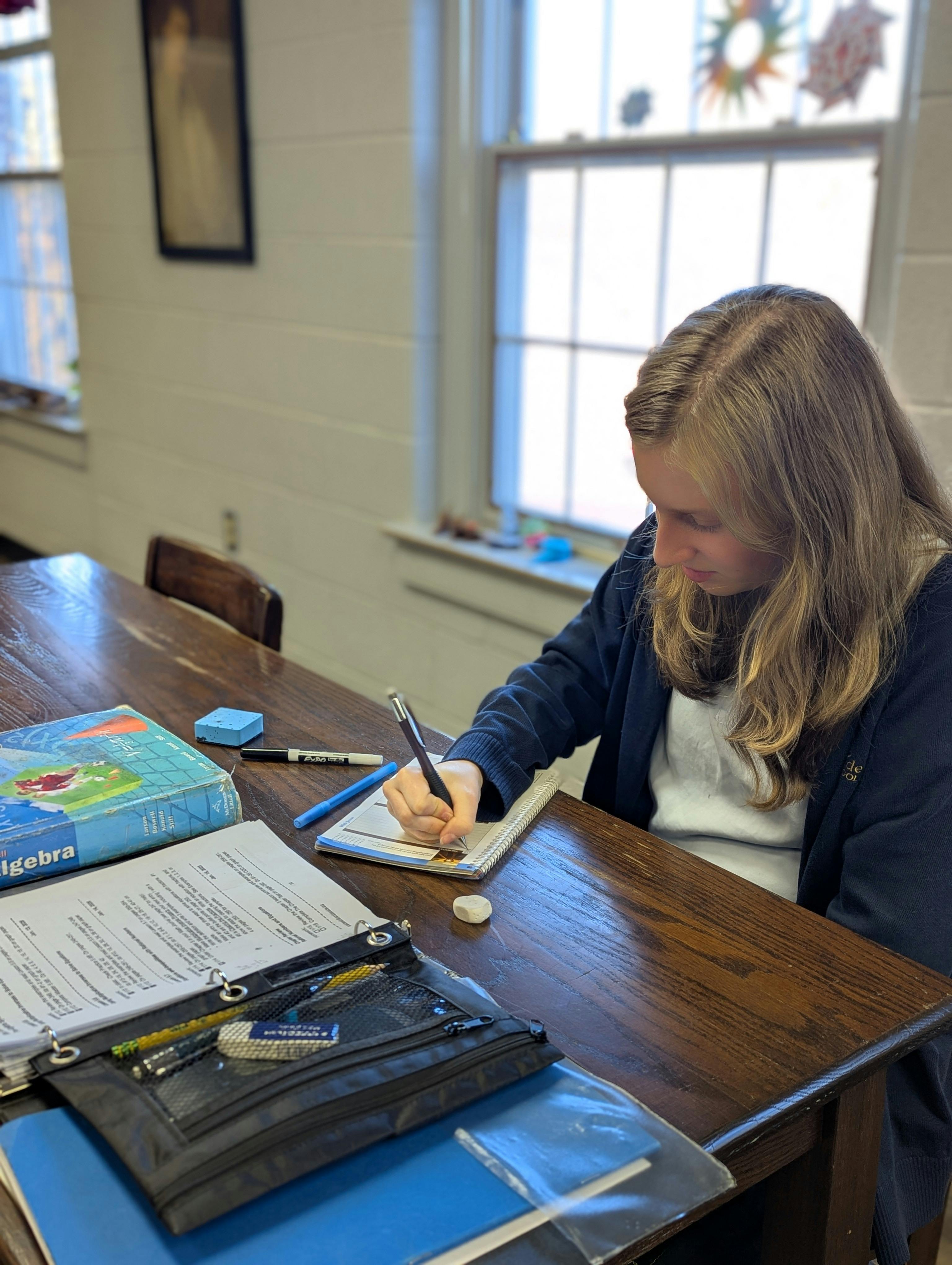 Child writing in a notebook at a desk with an algebra textbook