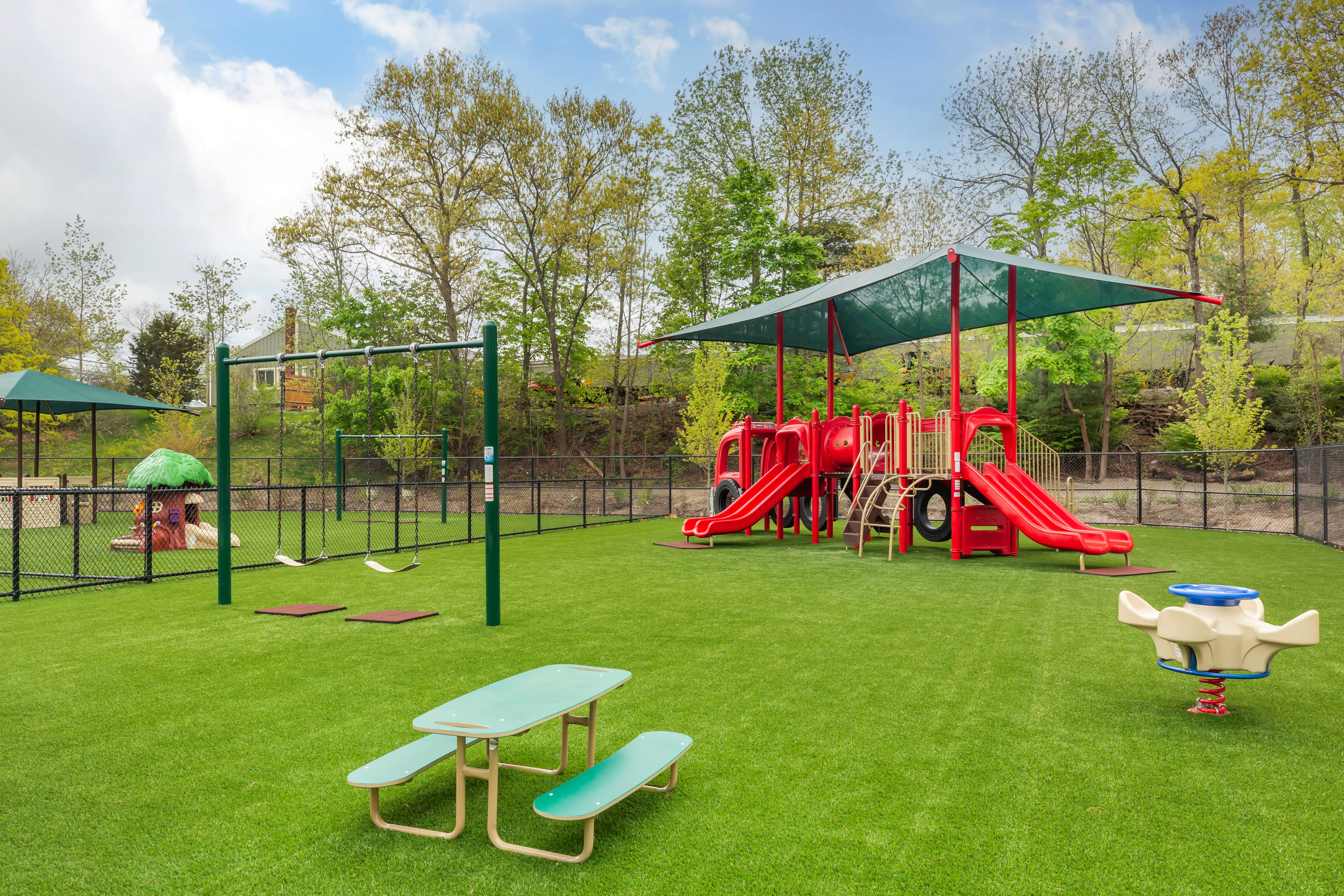 A fenced playground with a large red playset, swings, and a picnic table