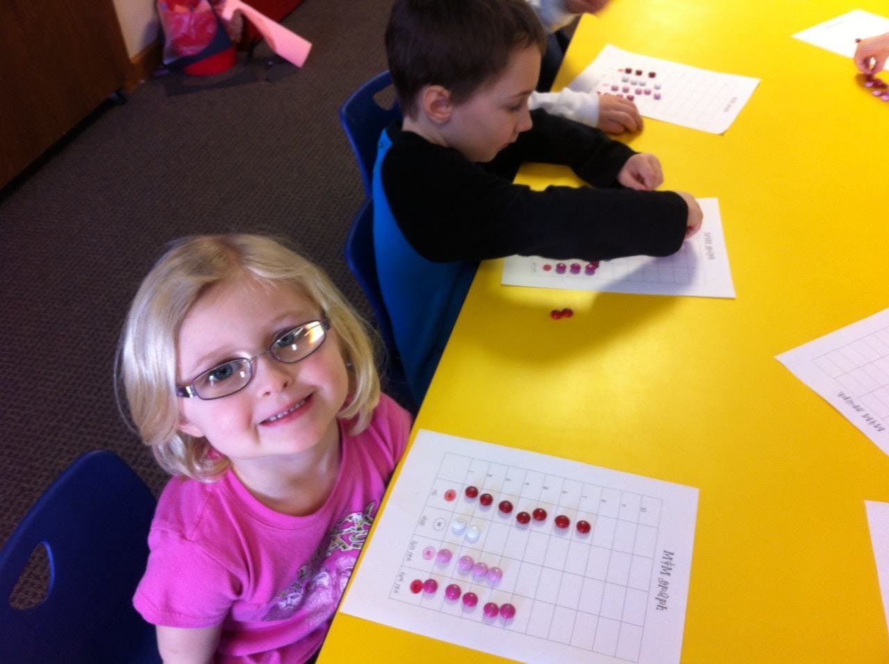 Child in glasses smiling at a table with another child working on a paper