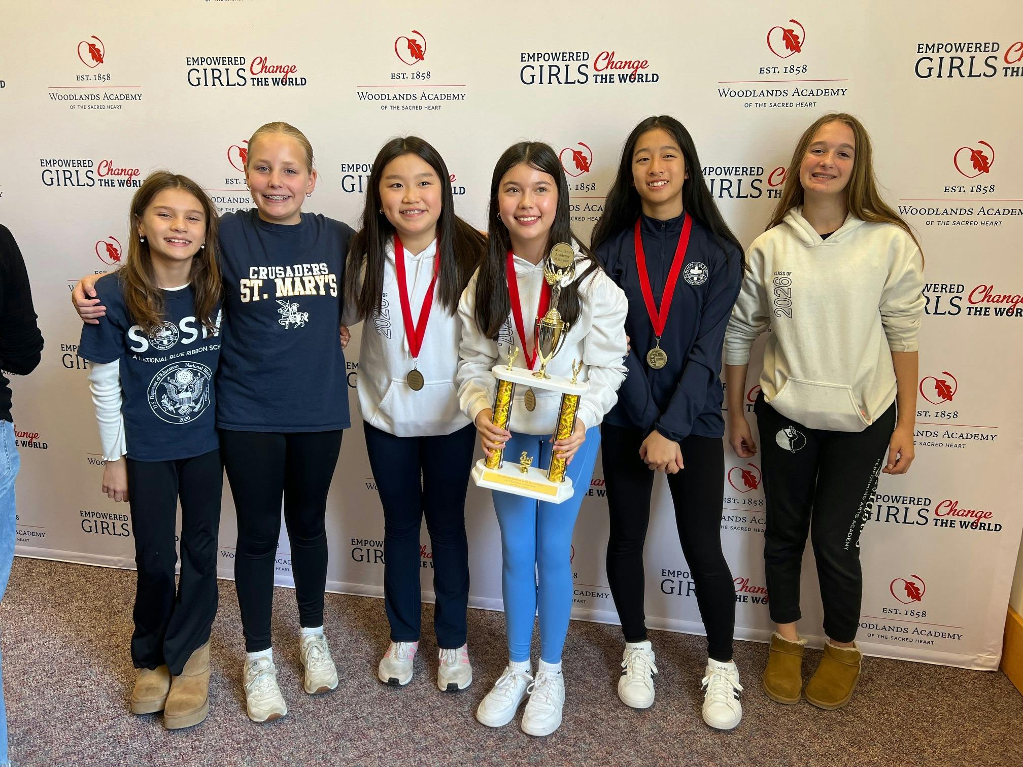 Six teens standing in a row, one holding a trophy, all wearing medallions