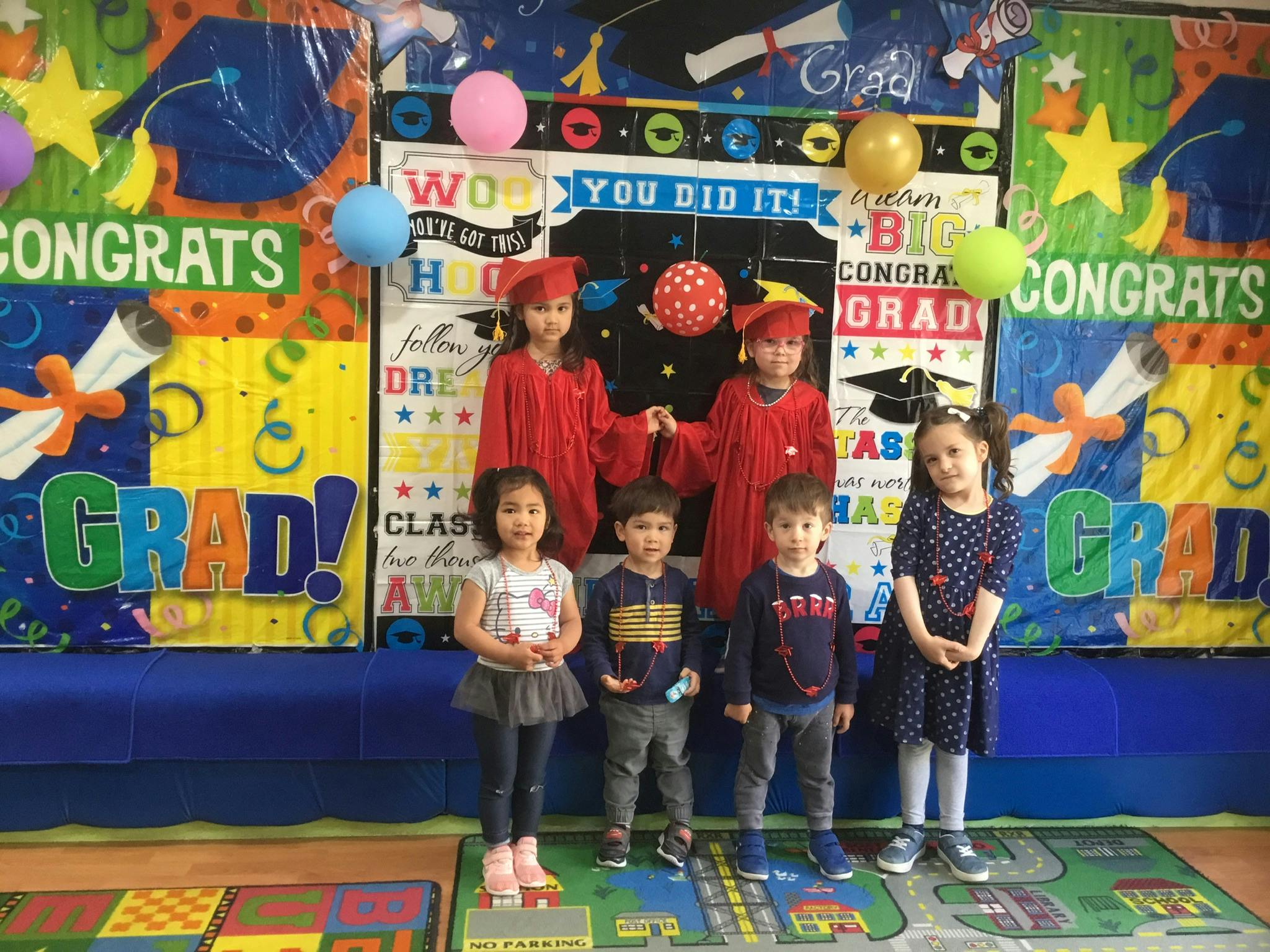 Children in graduation caps and gowns with other children in front of a banner