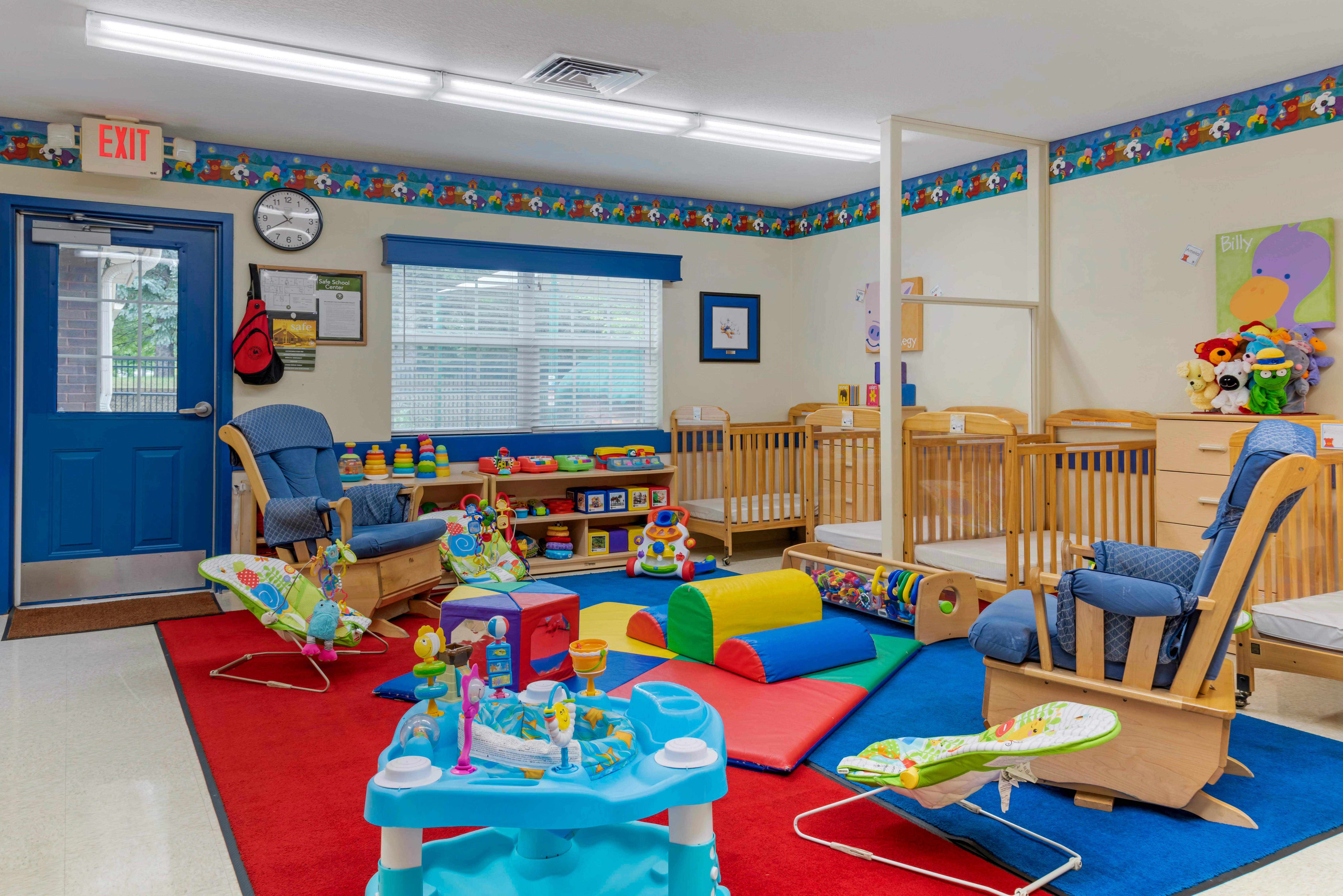 A childcare classroom with cribs, play mats, toys, and blue rocking chairs