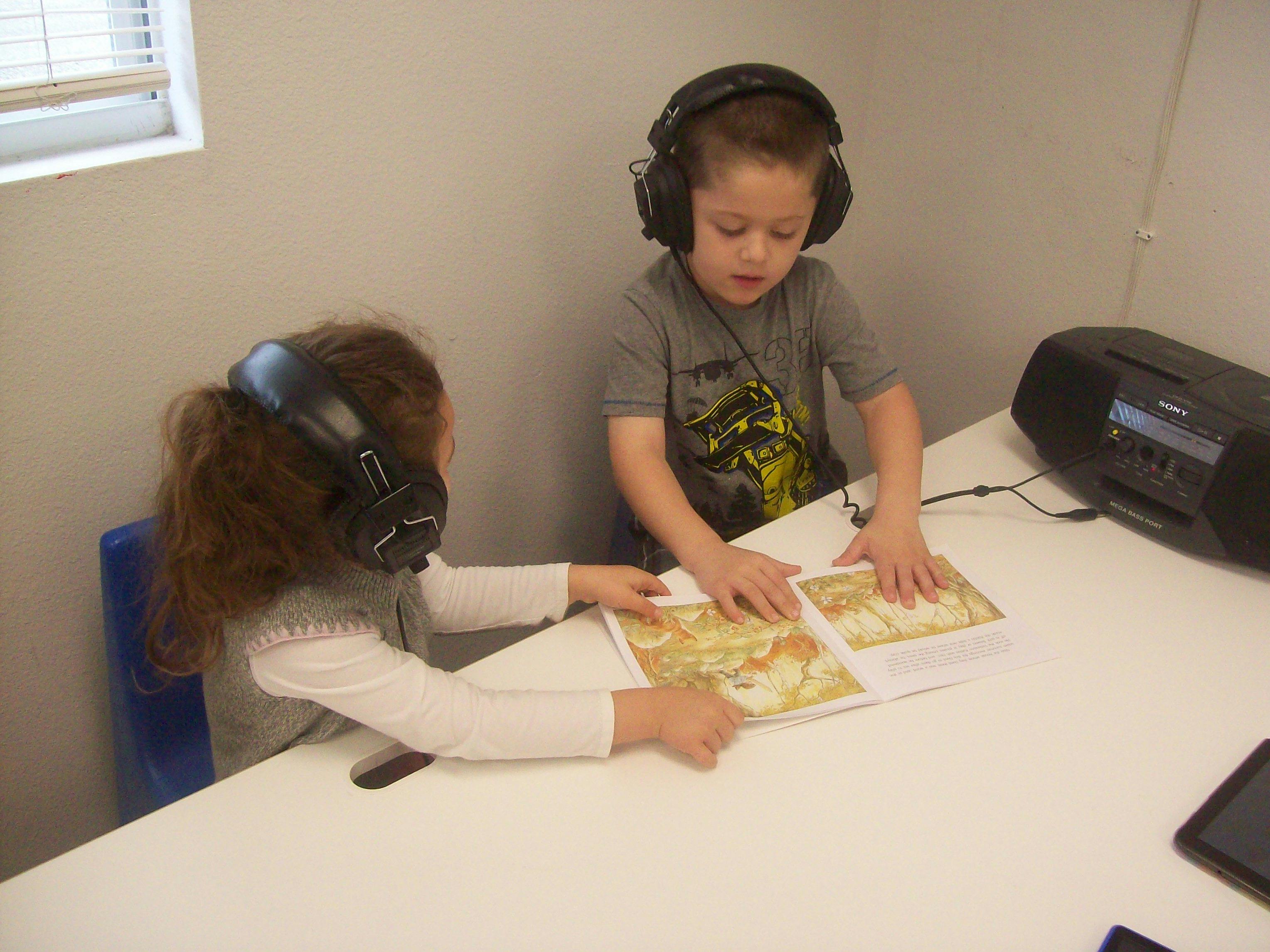 Two children wearing headphones reading a book together at a table