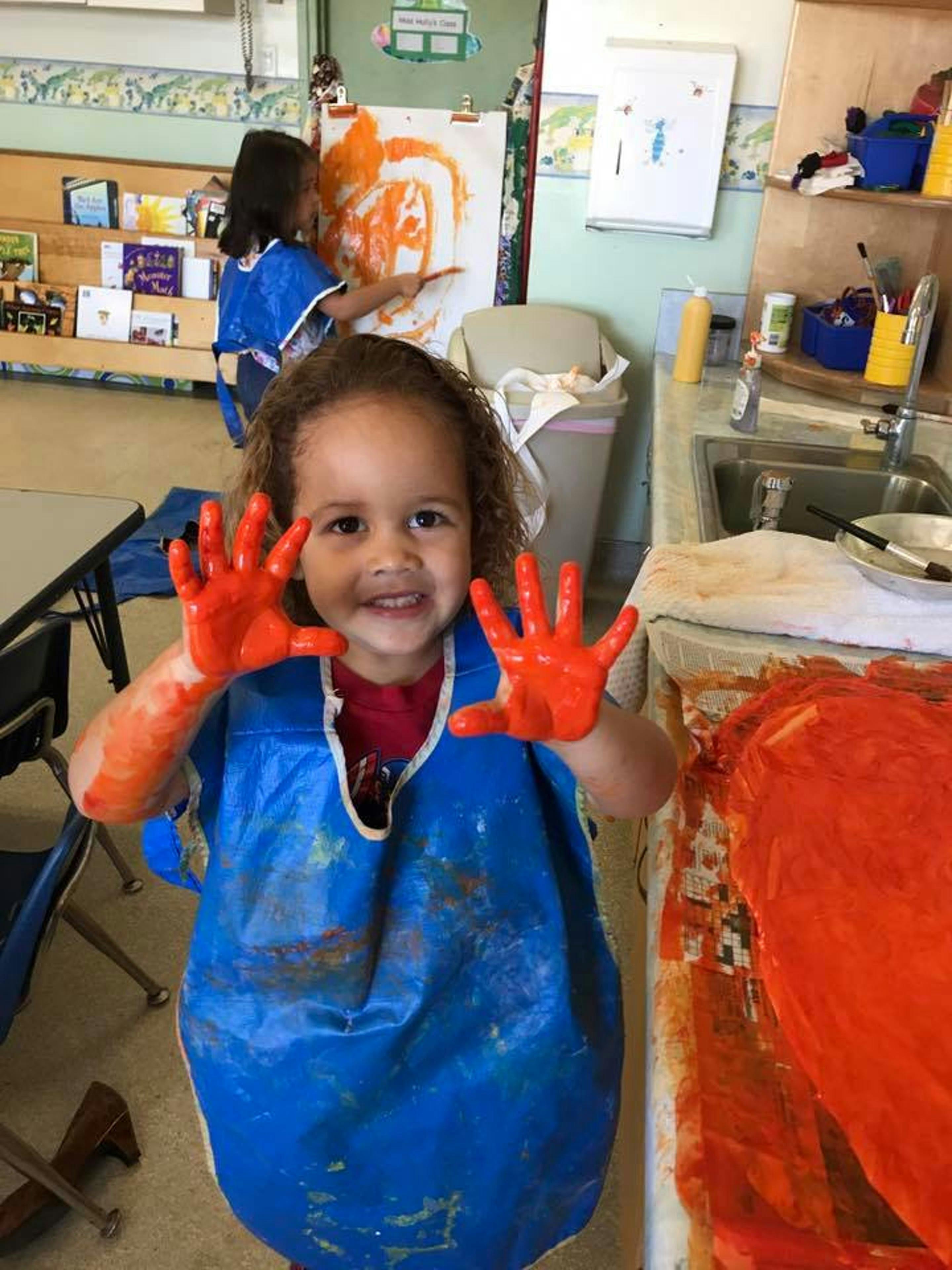 Child with orange paint on hands and arms, wearing a blue smock