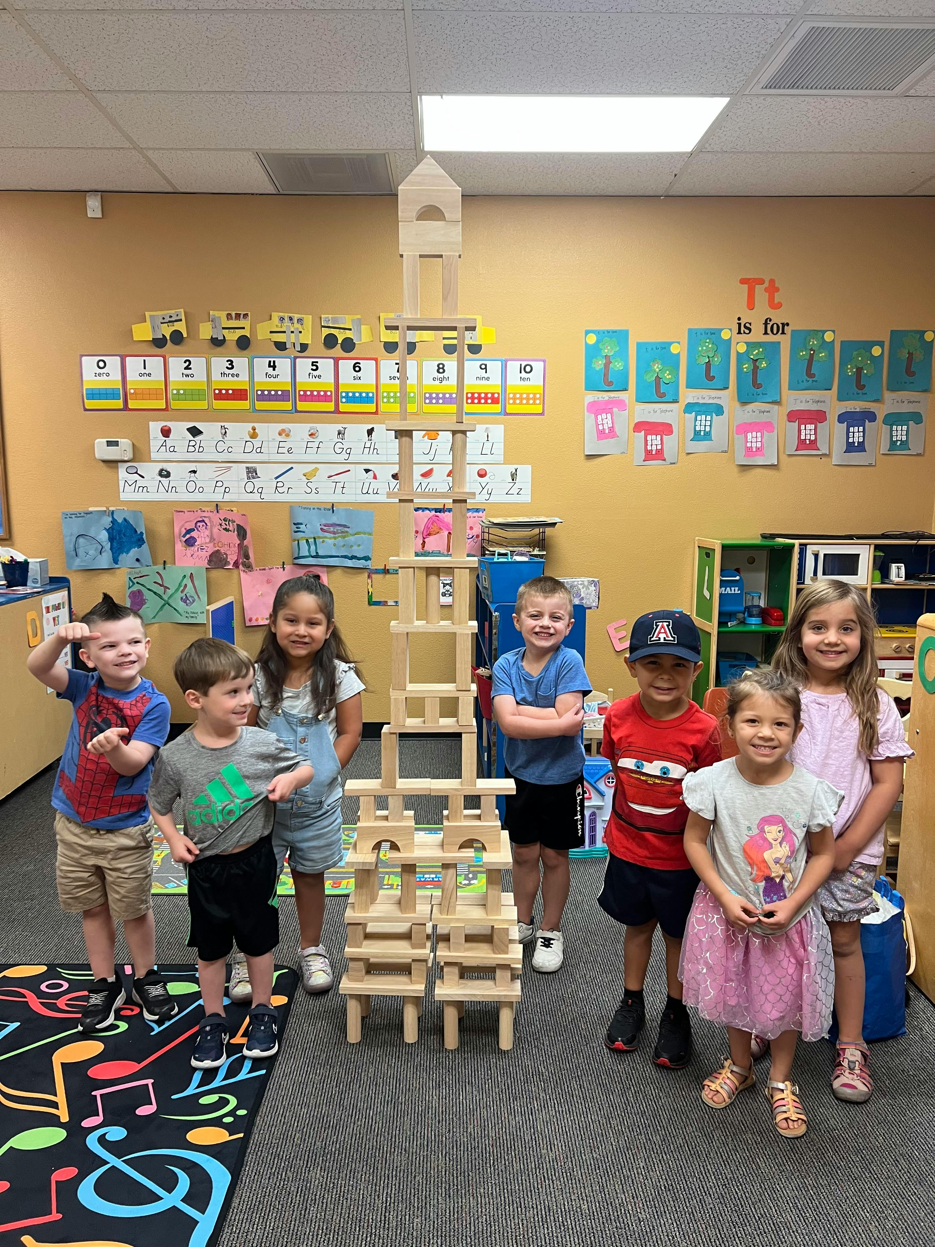 Group of children standing around a tall wooden block tower in a classroom