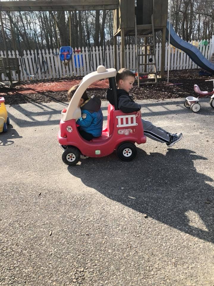 Two children in a red toy car on pavement