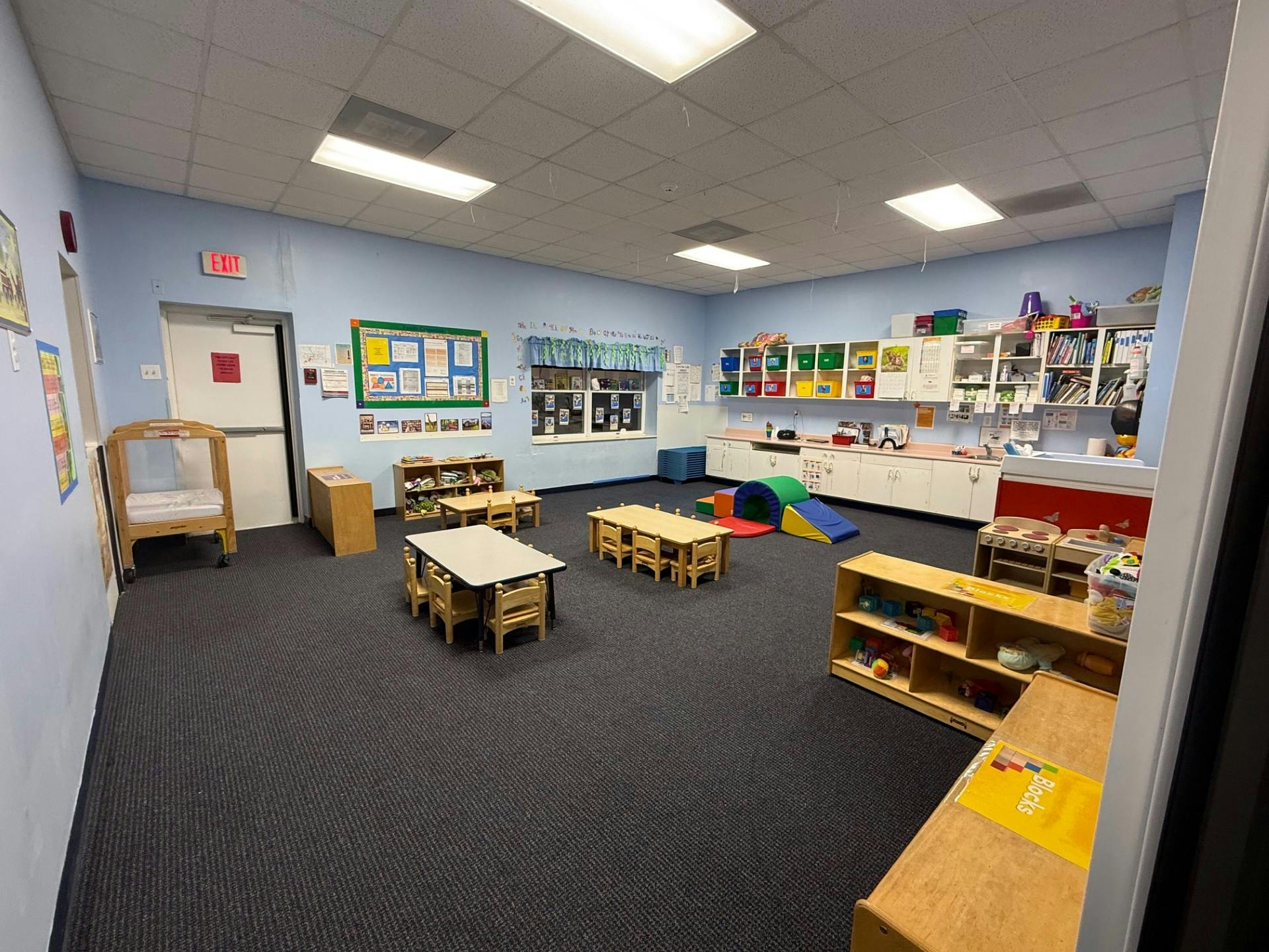 Classroom with small tables and chairs, play mats, and storage shelves