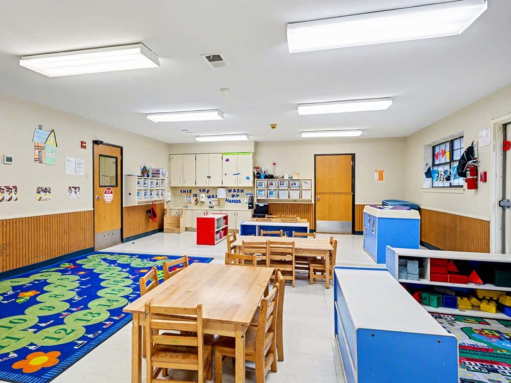 Classroom with small wooden tables and chairs, and a blue rug with numbers