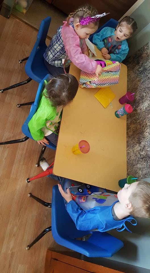 Three children at a table, one opening a colorful box