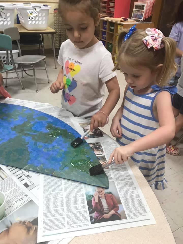 Two children painting a large blue and green cutout in a classroom