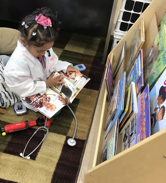 Child in a white coat reading a book next to a bookshelf