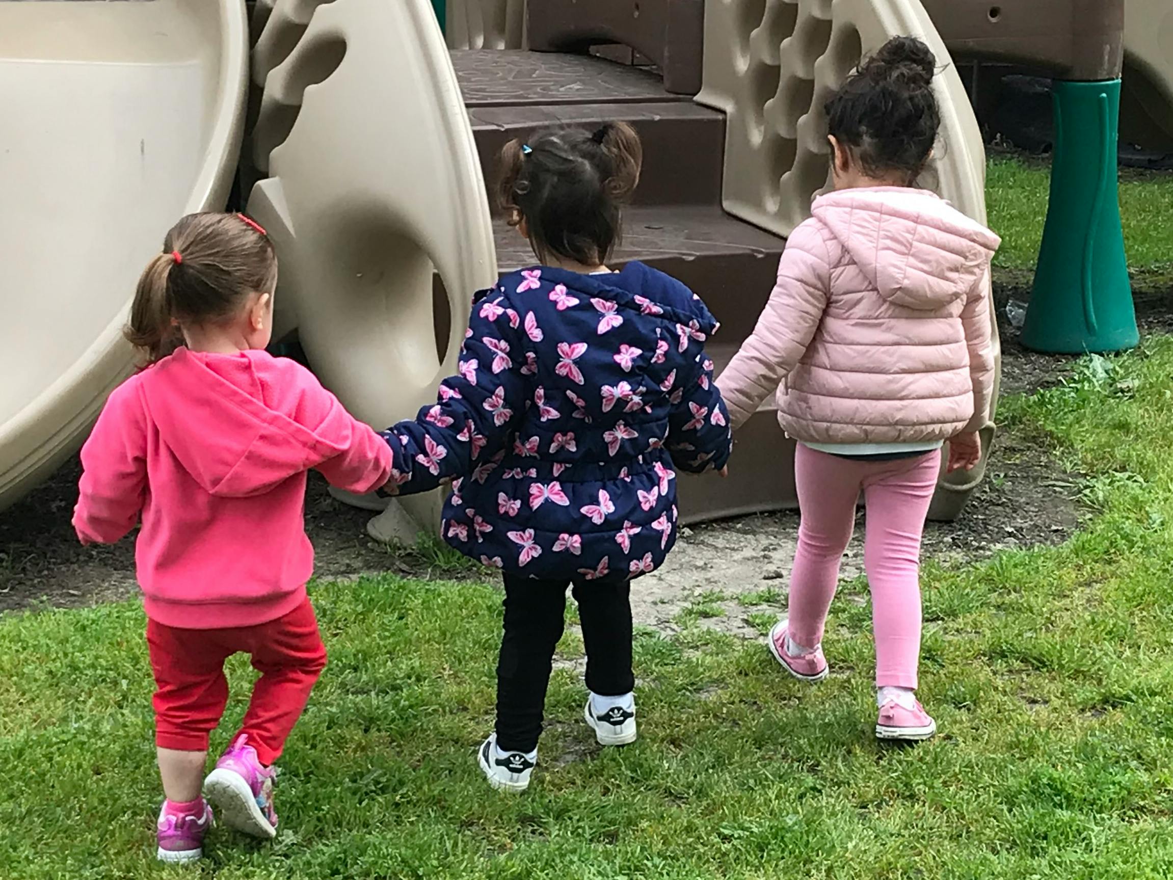 Three children holding hands walking on grass toward a playground slide