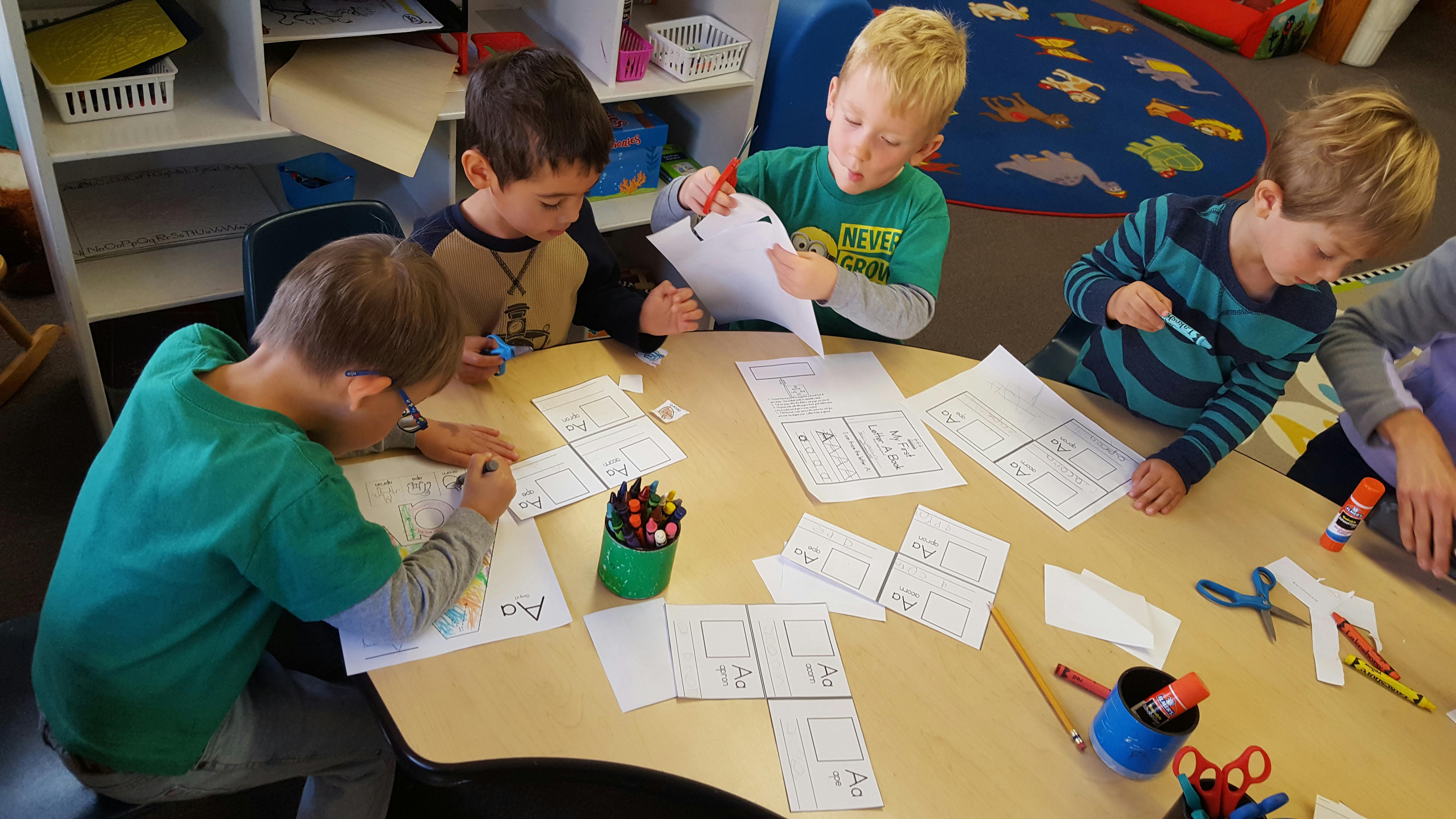 Four children drawing and cutting paper at a table in a classroom