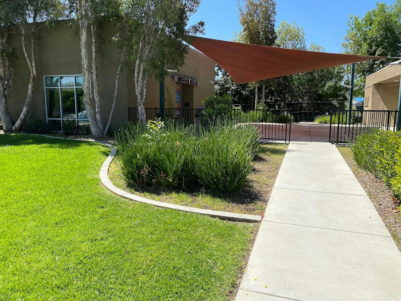 Building exterior with green lawn, bushes, and a concrete path leading to a shade sail