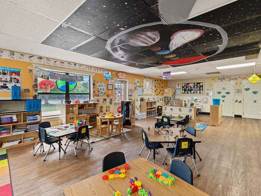 A classroom with small tables and chairs, shelves, and space-themed ceiling