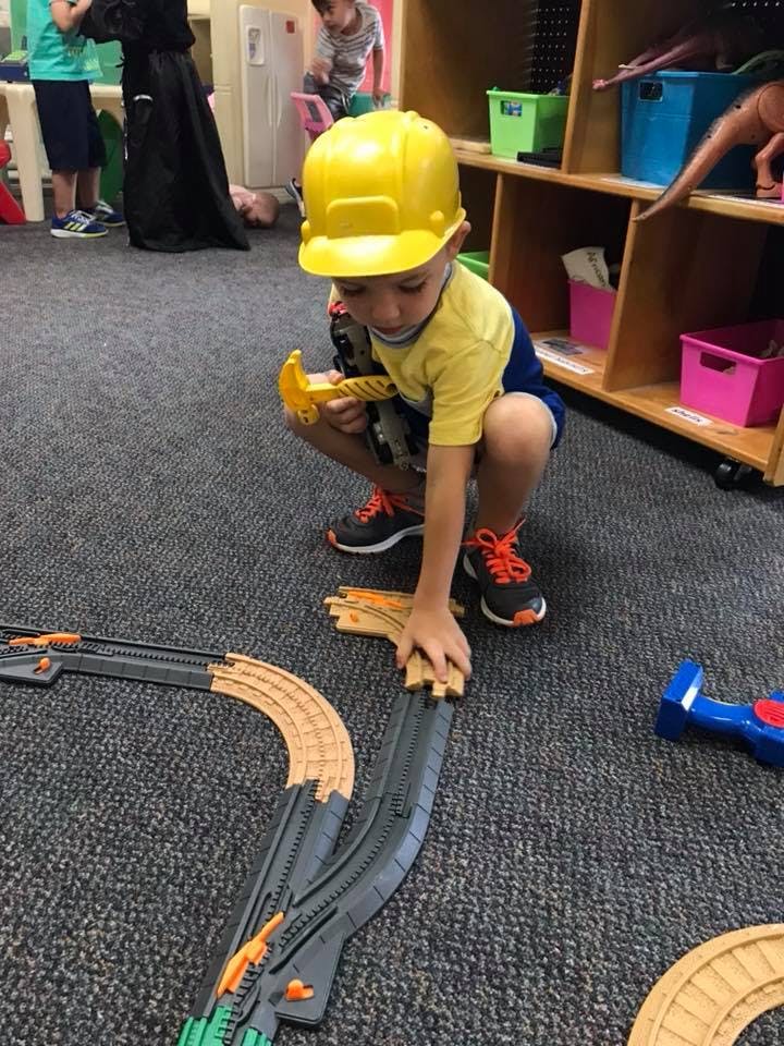 Child in a yellow hard hat playing with toy train tracks on the floor