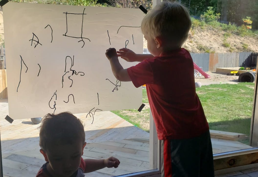 Child drawing on paper taped to a window, another child in foreground