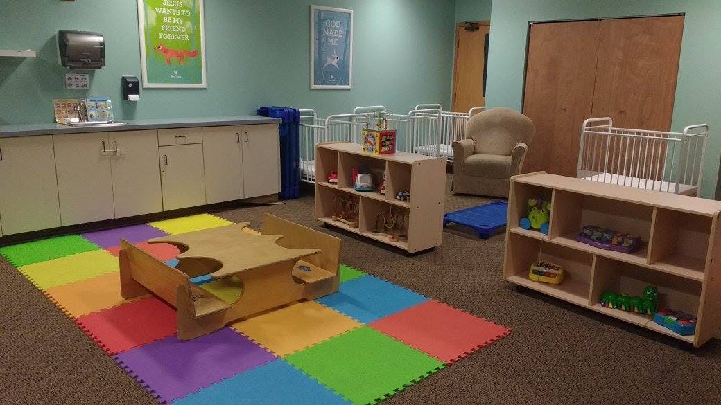 Playroom with colorful floor mats, small table, shelves, and cribs