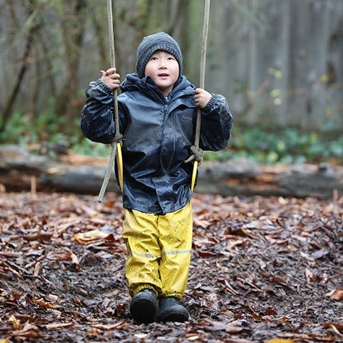 Child in a raincoat and rain pants holding onto a swing